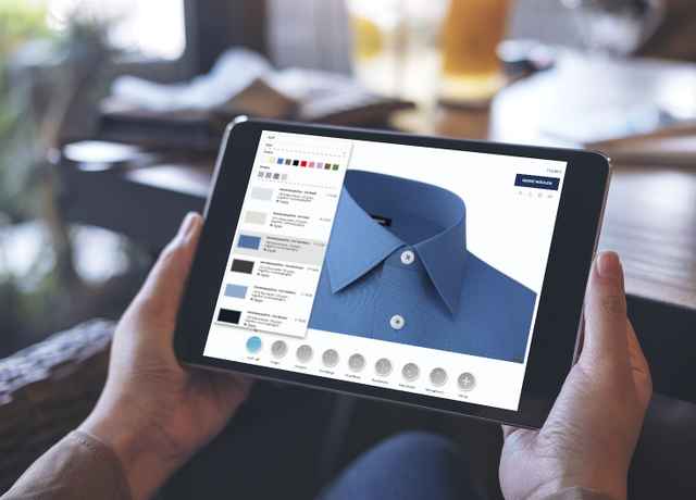 Person hält Tablet mit einer blauen Hemd-Design-App, die Stoff- und Knopfoptionen zeigt, in einem gemütlichen Café-Umfeld.