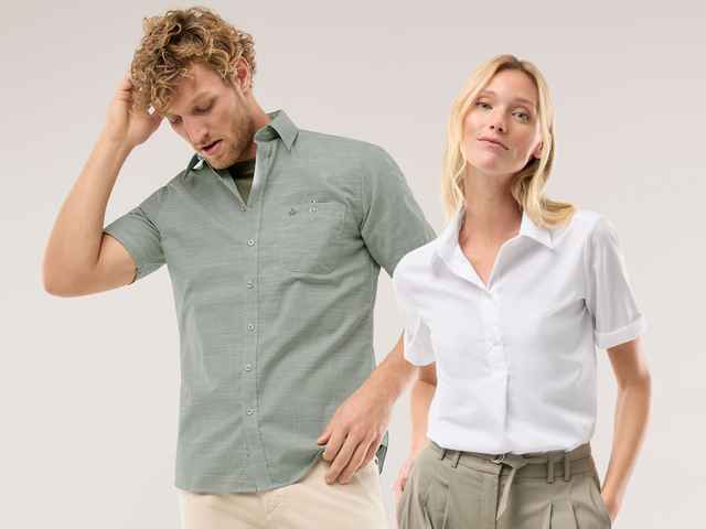 Ein Mann trägt ein Kurzarmhemd in Khaki, eine Frau trägt eine weiße Bluse und hält ihre Hände in den Hosentaschen.