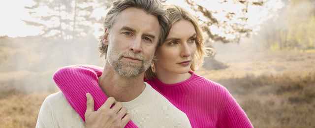 Ein Mann in cremefarbenem Pullover und eine Frau in pinkem Pullover stehen in einer herbstlichen Landschaft und blicken nachdenklich zur Seite.