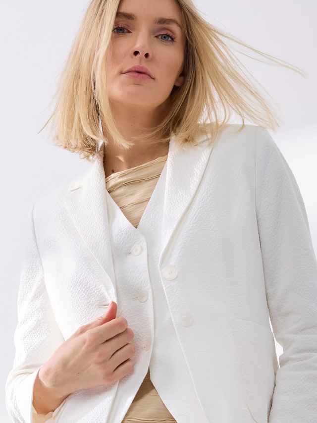 Frau mit blondem Haar trägt einen eleganten weißen Blazer und ein beiges Top, schaut selbstbewusst mit Wind in ihrem Haar.