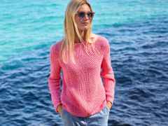 Frau mit Sonnenbrille, rosa Pullover und blauer Hose steht entspannt vor klarem, blauem Meerwasser.