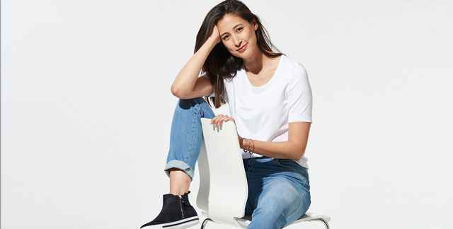 Frau in weißem T-Shirt und blauer Jeans sitzt auf einem Stuhl, lehnt den Kopf auf die Hand.