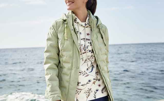 Frau in hellgrüner Jacke und floralem Hoodie steht lächelnd am Meer auf einem Pier.