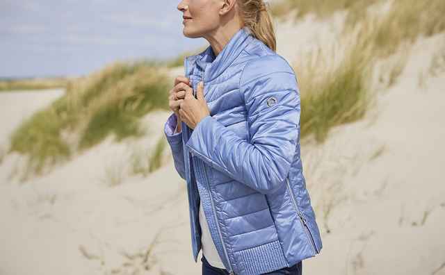 Glückliche Frau mit einer Steppjacke am Meer stehend.