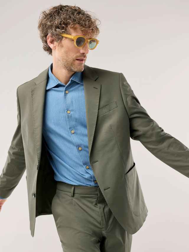 Ein Mann trägt einen olivgrünen Anzug mit hellblauem Hemd und gelber Sonnenbrille, was modern, leicht und lässig‑frisch wirkt.