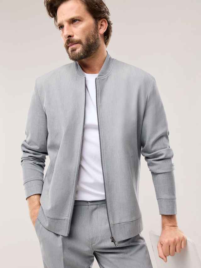 Ein Mann trägt ein hellgraues, modern geschnittenes Set aus Jacke und Hose über einem weißen Shirt, was einen klaren, frischen und ruhig‑lässigen Eindruck vermittelt.