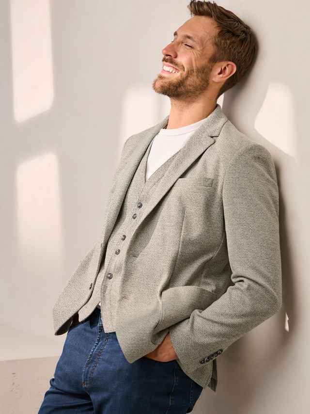 Ein Mann trägt eine hell strukturierte Jacke mit passender Weste zu Jeans und weißem Shirt, was modern‑lässig wirkt und eine warme, entspannte Stimmung ausstrahlt.