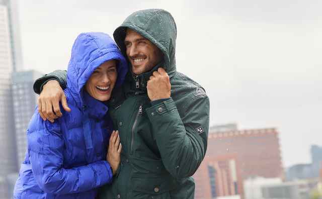 Ein lachendes Paar in blauen und grünen Jacken steht im Regen vor einer städtischen Skyline.