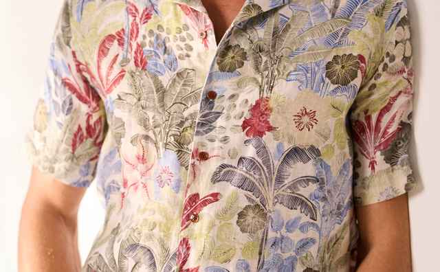 Mann in einem kurzärmeligen, floral bedruckten Hemd mit bunter Musterung und weißer Hose vor hellem Hintergrund.