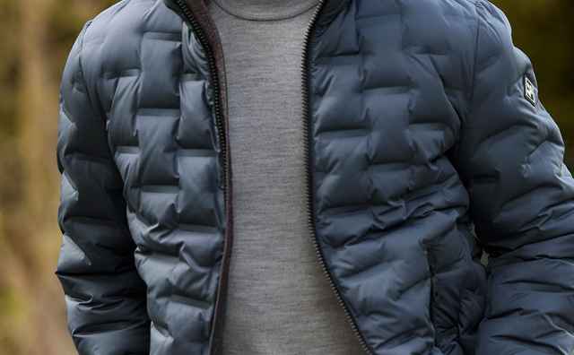 Mann mit grauen Haaren und Bart, trägt eine dunkelblaue Steppjacke und einen grauen Pullover, geht draußen entlang.