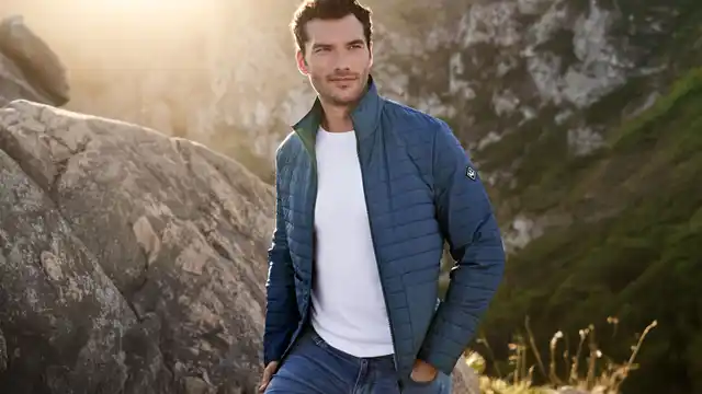 Ein Mann in blauer Jacke steht vor einer Felskulisse mit Sonnenlicht im Hintergrund, trägt ein weißes T-Shirt und blaue Jeans.