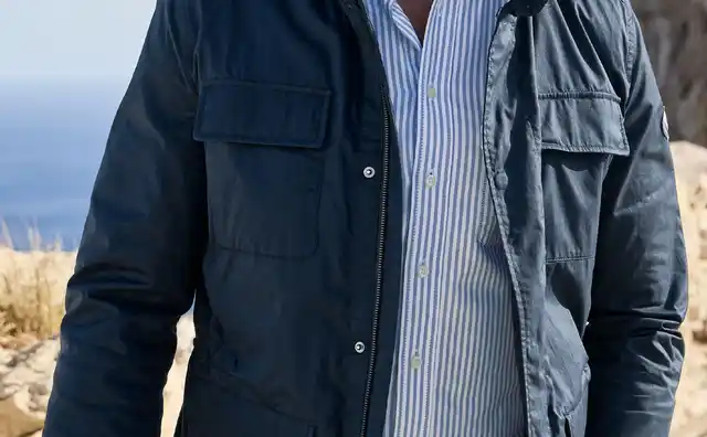 Ein Mann in einer blauen Jacke und einem gestreiften Hemd steht auf einer felsigen Küste mit Meer im Hintergrund.