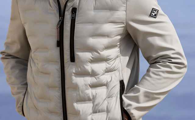 Ein Mann trägt eine beige Jacke mit Kapuze und steht vor einer Küstenkulisse. Er blickt zur Seite und hat die Hände in den Taschen.