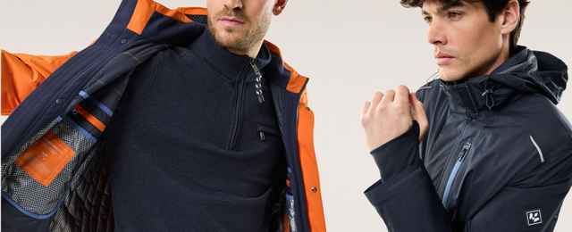 Zwei Männer in Winterbekleidung, einer in orange-blauer Jacke, der andere in dunkelblauer Jacke, posieren vor neutralem Hintergrund.