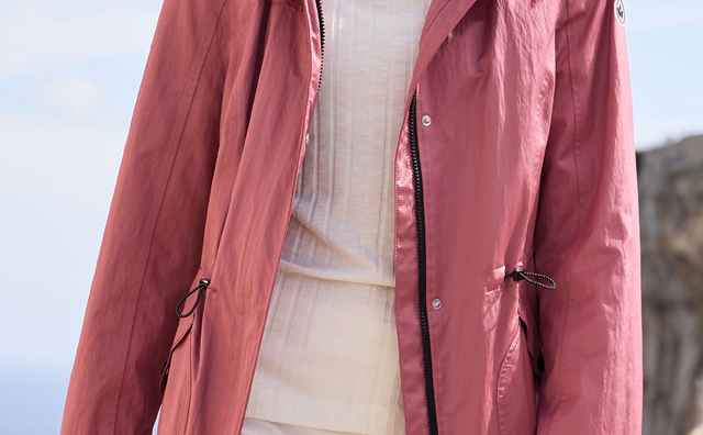 Frau in rosa Windjacke und weißer Hose steht im Freien, windgepeitschte Haare, mit Meer und Felshintergrund.