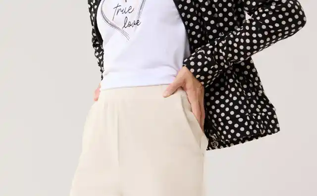 Eine Frau trägt eine schwarz‑weiß gepunktete Jacke über einem weißen T‑Shirt mit Herzmotiv und heller, weiter Hose. Der Look wirkt modern, lässig und leicht elegant.