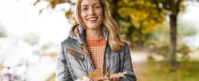 Frau in Herbstkleidung lächelt, hält Herbstblätter in der Hand, im Hintergrund herbstliche Bäume und unscharfe Landschaft.