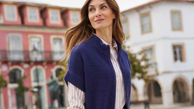 Frau in blauer Strickweste steht lächelnd vor einem roten Haus.