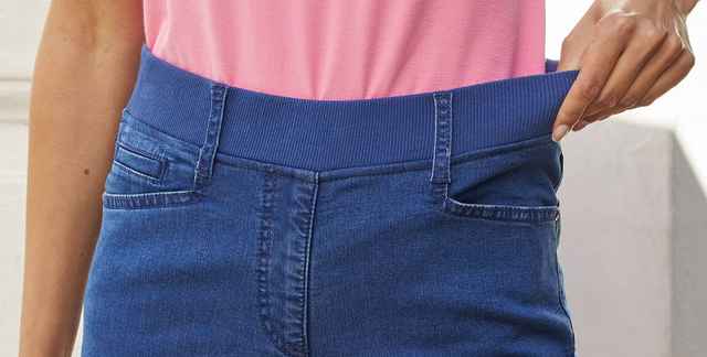 Eine Person trägt eine blaue Jeans mit elastischem Bund und ein rosa Oberteil, während sie den Bund der Hose mit der Hand zieht.