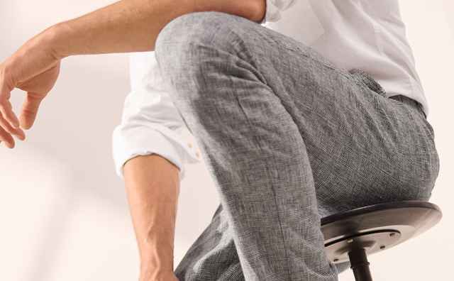 Mann in weißem Hemd und grauer Hose sitzt auf einem Hocker, trägt weiße Sportschuhe und schaut ernst in die Kamera.