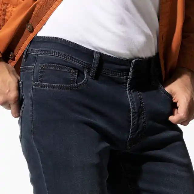 Person trägt dunkelblaue Jeans, weißes T-Shirt und orange Jacke, mit den Händen in den Hosentaschen.