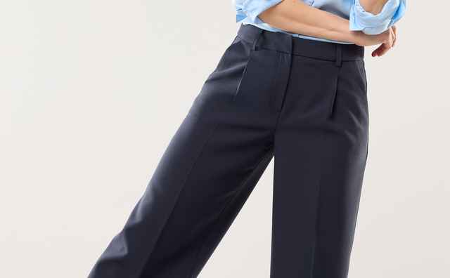Frau in blauer Bluse, dunkler Hose und hellblauen Sneakern steht lächelnd und locker in einer Pose, die Entspannung und Stil ausstrahlt.