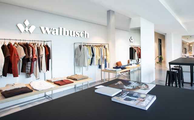 Innenansicht eines Bekleidungsgeschäfts von Walbusch mit hängenden Kleidungsstücken und Auslagen auf Tischen.