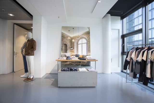 Moderne Boutique mit Kleidung auf einem Tisch, einer Schaufensterpuppe in Casual-Outfit und einem Kleiderständer; große Fenster im Hintergrund.