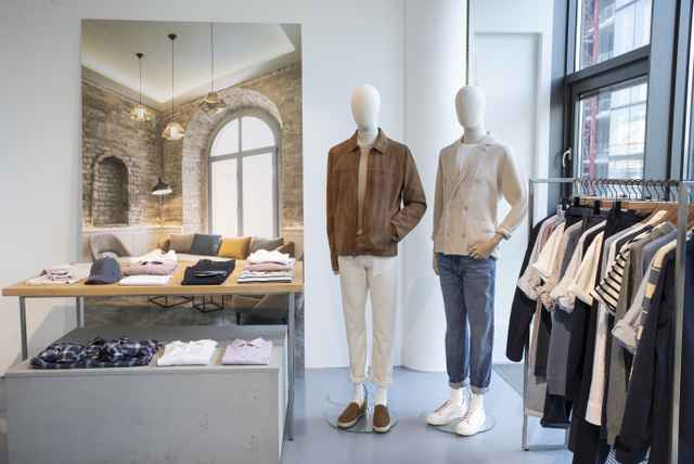Schaufenster in einem Bekleidungsgeschäft mit zwei Schaufensterpuppen, einem Tisch mit gefalteten Kleidungsstücken und einem Kleiderständer.