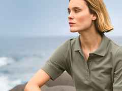 Frau mit blondem Haar und olivgrünem Poloshirt blickt nachdenklich aufs Meer. Im Hintergrund sind Wellen und ein bewölkter Himmel zu sehen.