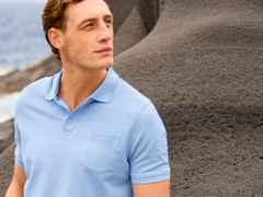 Mann in hellblauem Poloshirt steht vor einer dunklen Felsformation und blickt in die Ferne. Im Hintergrund ist teils das Meer zu sehen.