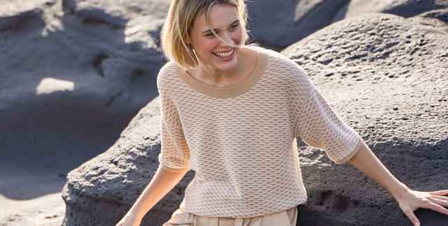 Lächelnde Frau in beigefarbenem Pullover sitzt auf dunklen Felsen im Freien.
