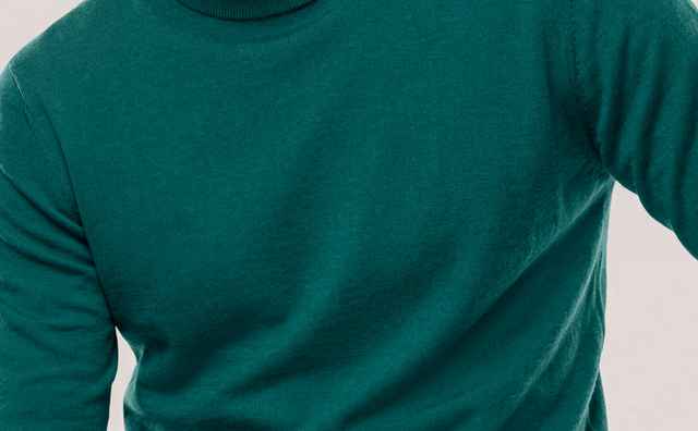 Mann mit braunen Haaren trägt einen grünen Rollkragenpullover und blaue Jeans, sitzt vor neutralem Hintergrund und schaut ernst.