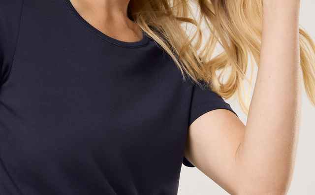 Frau mit blondem Haar trägt ein dunkelblaues T-Shirt und lächelt leicht, während sie ihre Hand durch das Haar fährt.