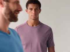 Zwei Männer im Vordergrund, einer lächelt in einem blauen T-Shirt, der andere ernst im lila T-Shirt vor grauem Hintergrund.