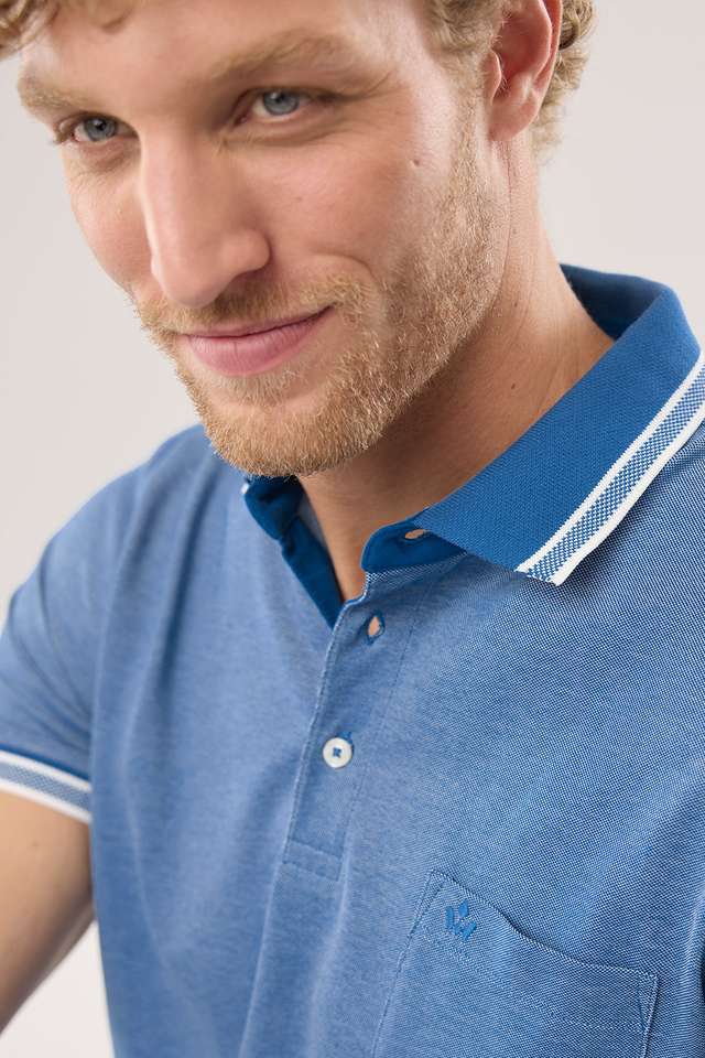 Mann in blauem Poloshirt und hellblauen Hosen, kniet auf dem Boden und blickt in die Kamera vor grauem Hintergrund. Angezeigt beim Hover