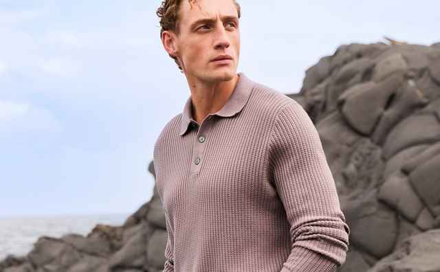 Ein Mann in einem beigefarbenen Pullover steht auf Felsen am Meer, schaut nach rechts. Der Himmel ist teilweise bewölkt.