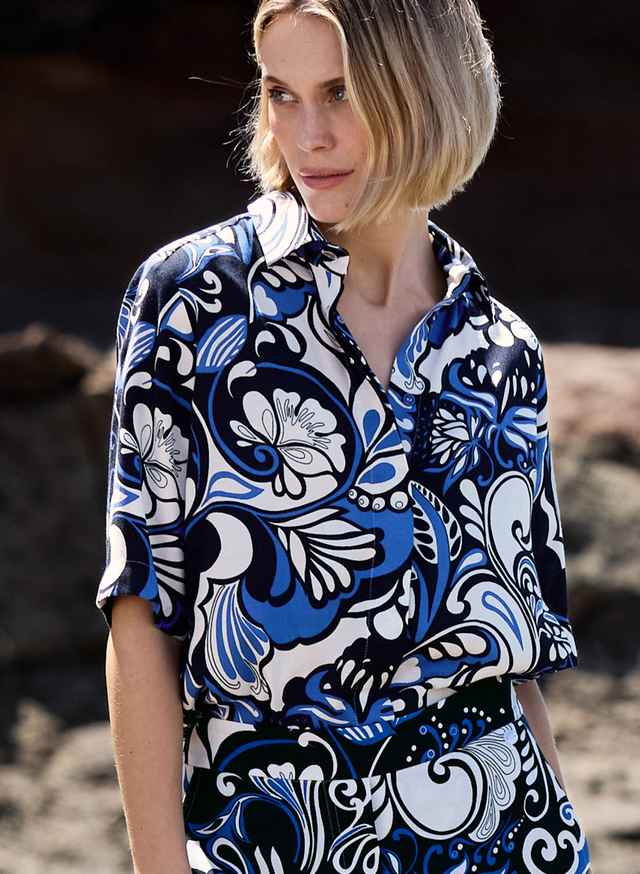 Bluse mit auffälligem floralen Muster in Blau, Schwarz und Weiß sowie klassischem Kragen und kurzen Ärmeln. Der Stil wirkt farbenfroh und verspielt.