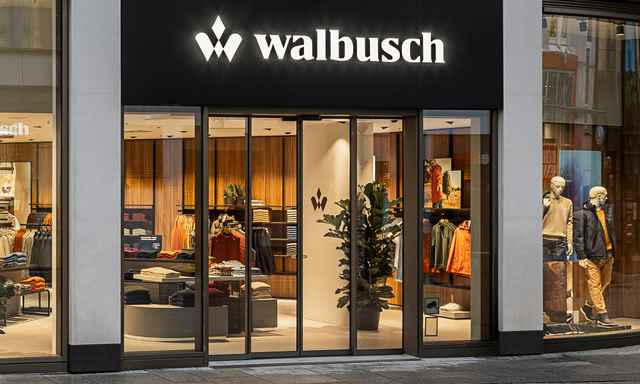 Eingang eines Walbusch-Geschäftes mit Schaufensterpuppen, Kleidung und Pflanzen dekoriert, beleuchtet von einem Leuchtschild mit dem Walbusch-Logo.
