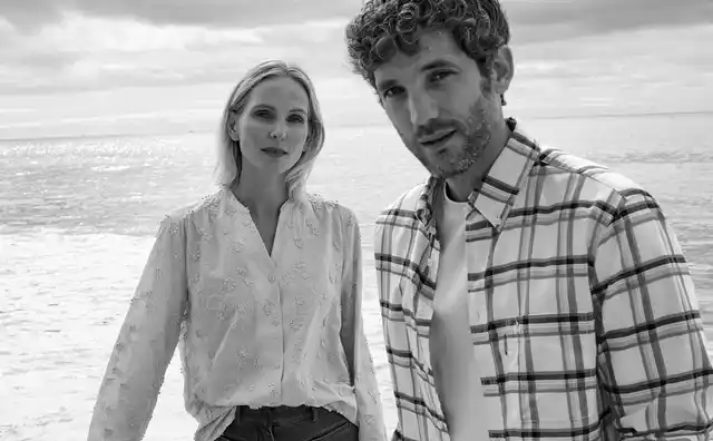 Ein Frau und ein Mann stehen am Strand. Sie trägt eine bestickte Bluse und er ein offenes kariertes Hemd mit einem weißen Shirt darunter.