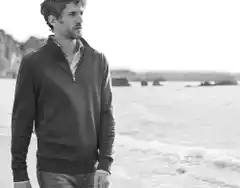 Ein Mann geht am Strand entlang und trägt einen Troyer, darunter ein gestreiftes Hemd und dazu eine Jeans.