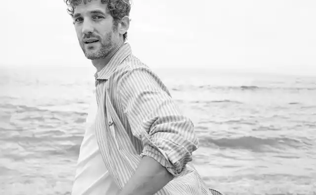 Ein Mann steht am Strand und trägt ein offenes Hemd mit einem Shirt darunter.