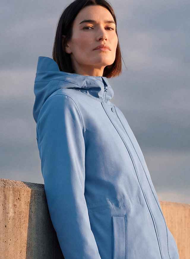 Frau in blauer Jacke mit Kapuze steht im Freien, lehnt an einer Mauer, Blick in die Ferne, bei bewölktem Himmel.