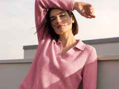 Frau in einem rosa Pullover, die draußen steht, mit einem Arm über dem Kopf, der Wind weht leicht durch ihr Haar, blauer Himmel im Hintergrund.