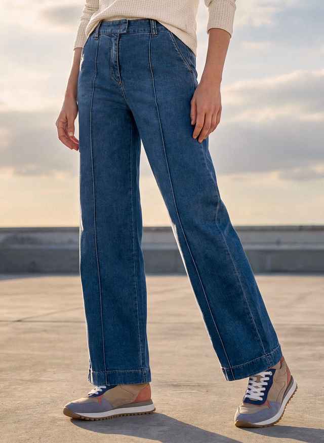 Person in blauer Jeans und Turnschuhen steht auf einer Betonfläche, mit Himmel im Hintergrund.