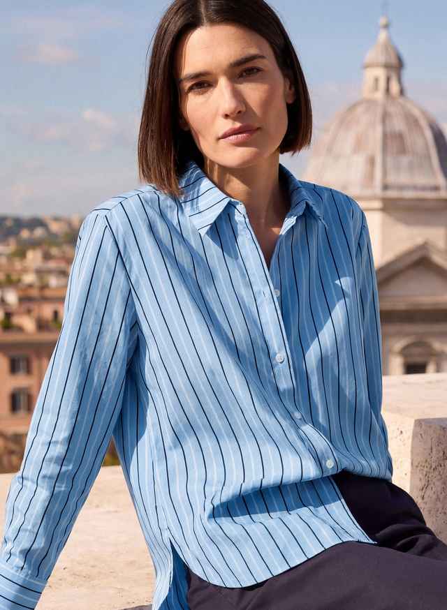 Frau in blau gestreifter Bluse sitzt vor einer Stadtlandschaft mit historischer Architektur im Hintergrund.