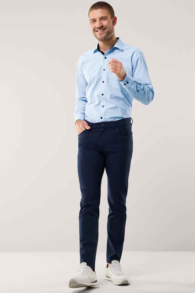 Mann in hellblauem Hemd und dunkelblauer Hose lächelt, steht in neutralem Hintergrund, eine Hand in der Hosentasche, trägt weiße Sneaker.