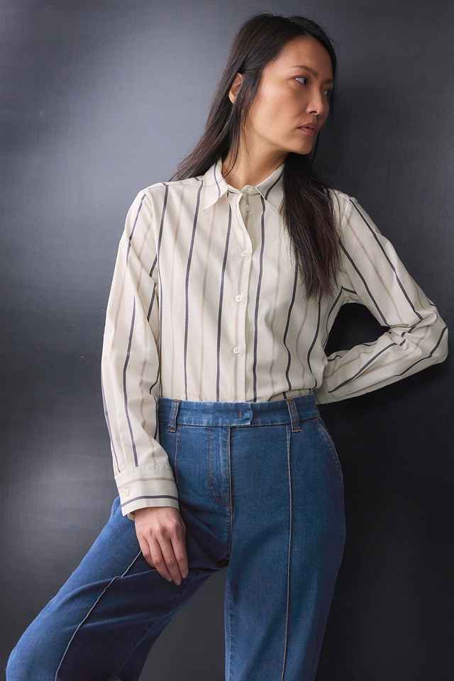 Frau in gestreiftem Hemd und Jeans steht vor dunklem Hintergrund und blickt nach links.