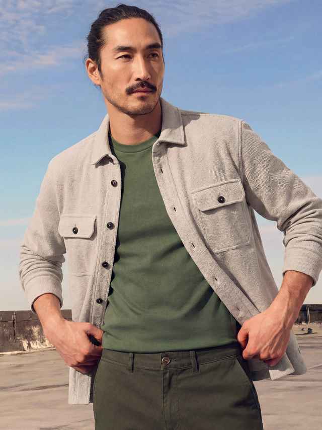 Mann mit dunklem Haar, trägt ein grünes T-Shirt und eine beige Jacke, steht unter blauem Himmel.