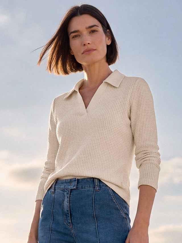 Frau mit Bob-Frisur in einem cremefarbenen Pullover und blauer Jeans, steht draußen bei Tageslicht mit einem klaren Himmel im Hintergrund.